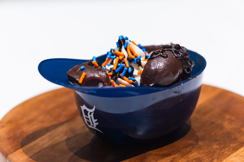 A chocolate ice cream sundae in a blue baseball cap-shaped bowl, topped with orange and blue sprinkles and chocolate drizzle.