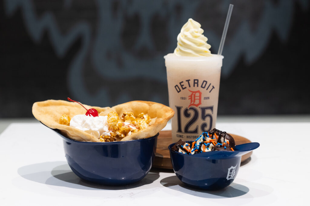 A Detroit Tigers-themed dessert spread featuring a popcorn sundae in a blue baseball cap bowl, a chocolate sundae with sprinkles, and a milkshake topped with whipped cream in a branded cup.