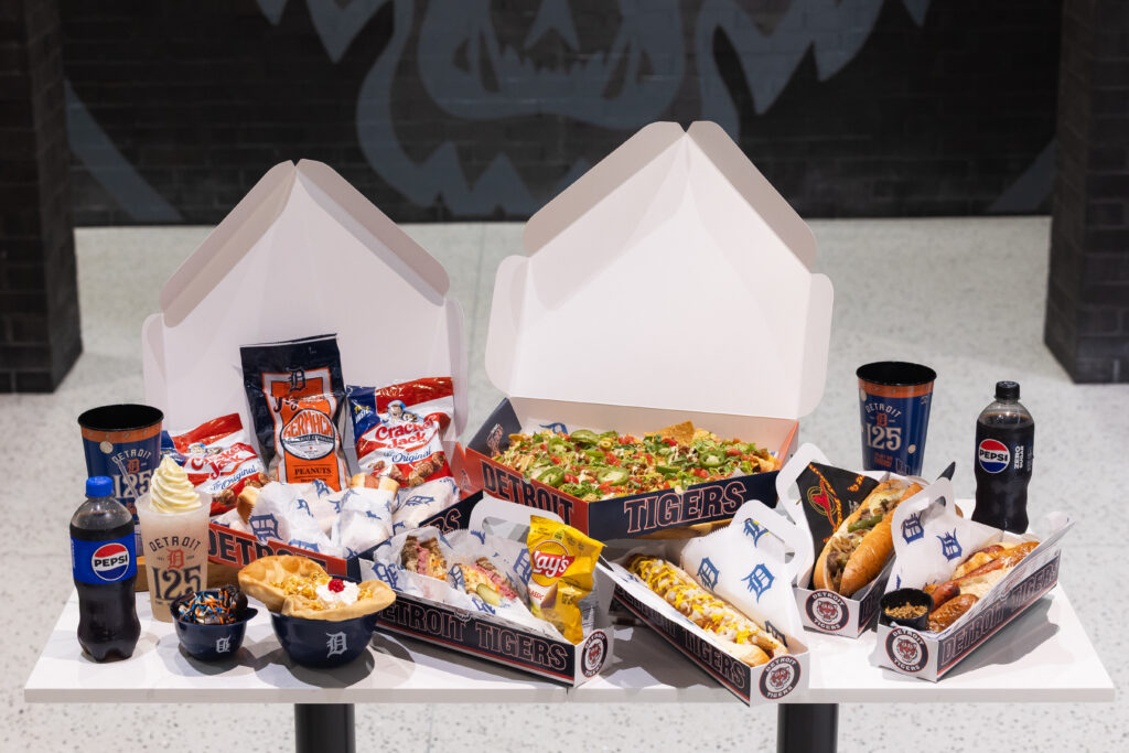 A variety of Detroit Tigers-themed food and drinks displayed on a table, including nachos, hot dogs, peanuts, and soda.